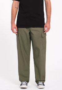 Pantalons cargo verts avec une coupe ample, deux poches latérales et un tissu robuste. Portés avec un t-shirt noir et des baskets noires avec des semelles blanches.