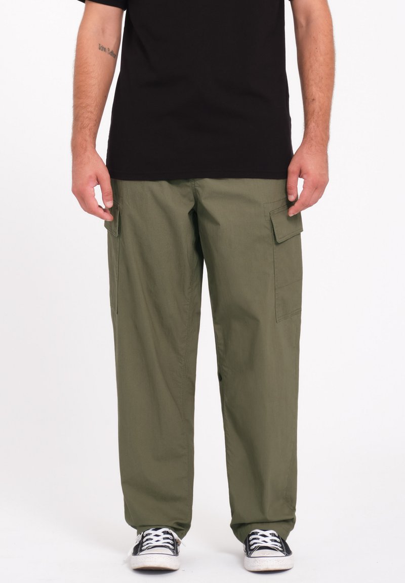 Pantalons cargo verts avec une coupe ample, deux poches latérales et un tissu robuste. Portés avec un t-shirt noir et des baskets noires avec des semelles blanches.