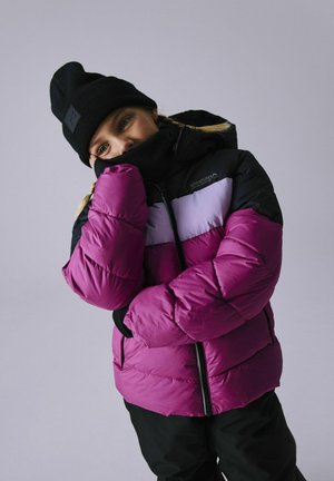 Young person wearing a black beanie and purple-black winter jacket, covering part of their face with a hand against a plain light background.