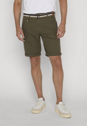 Mężczyzna w oliwkowozielonych podwiniętych szortach, pasku w paski, białej koszulce i białych trampkach stoi na jednolitym tle.