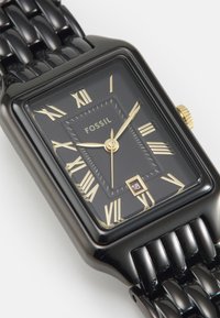 Svart rektangulär armbandsur med guldfärgade romerska siffror och visare, ett datumfönster och ett texturerat armband i svart metall.