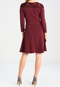 Mujer vistiendo un vestido burdeos a la altura de la rodilla con detalles de encaje en la parte superior de la espalda y mangas tres cuartos, combinado con tacones altos grises.