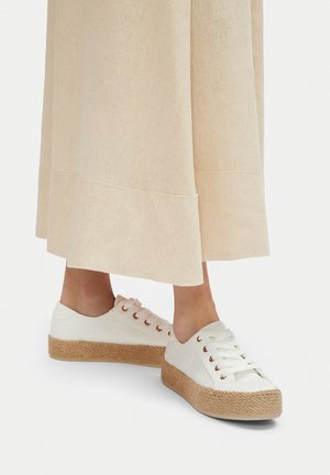 Białe sneakersy na platformie z teksturowaną cholewką, sznurowane, z miedzianoróżowymi przelotkami, zestawione z beżową, lejącą się spódnicą z obszytym dołem.