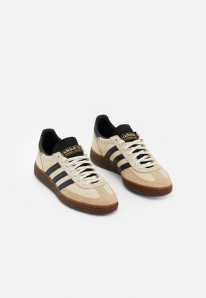 Par beige Adidas Handball Spezial sneakers med sorte striber, brune såler, hvide snørebånd og sorte hælkant på hvid baggrund.