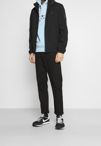 Černá lehká bunda s kapucí, v kombinaci se světle modrou mikinou a černými kalhotami. Doplněno sportovními černo-bílými teniskami.