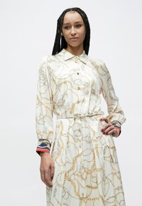 Krekls ar garām piedurknēm krēmkrāsas audumā ar zelta ķēdes rakstu. Aprīkots ar apkakli, pogājamu priekšpusi un atbilstošu jostu ar krāsainiem aproču akcentiem.