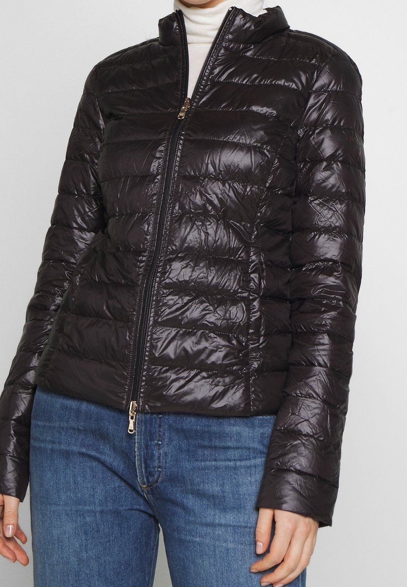 Veste matelassée noire avec fermeture éclair à l'avant, portée sur un col roulé blanc et un jean bleu, montrant le torse et les bras d'une personne.