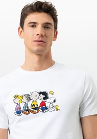 Jongeman draagt een wit T-shirt met een kleurraf grafiek van Peanuts-personages die in een rij op de voorkant staan.