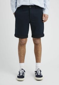 Shorts bleu marine en mélange de coton, avec des ourlets retroussés, des poches avant et une fermeture à bouton. Portés avec des chaussettes blanches et des baskets bleu marine.