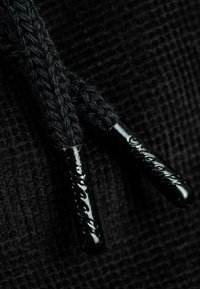 Nahaufnahme eines schwarzen Strickstoffs mit zwei schwarzen geflochtenen Schnüren, die an ihren Enden glänzende, geprägte Kunststoffspitzen aufweisen.