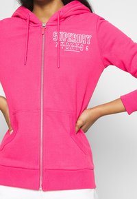 Frau in einem leuchtend pinken Hoodie mit Reißverschluss, auf dem der Text "Superdry Tokyo 76 Athletic Department" steht, mit den Händen in den vorderen Taschen.