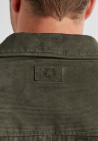 Eine olivgrüne Cordjacke mit strukturiertem Finish, ausgestattet mit einem rechteckigen Stoffetikett, das auf den Rücken genäht ist und ein Logo-Detail zeigt.