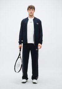 Tuta sportiva blu navy con giacca con zip e pantaloni con strisce laterali bianche. Indossata con una maglietta bianca e una racchetta da tennis.