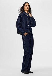 Femme debout portant une veste en denim bleu foncé et un jean assorti avec des clous argentés, une main dans la poche, sur un fond clair uni.