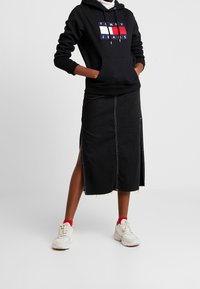 Falda de denim negra con aberturas laterales, dobladillo sin terminar y costuras blancas. Combinada con una sudadera negra que tiene un logotipo multicolor y zapatillas blancas.