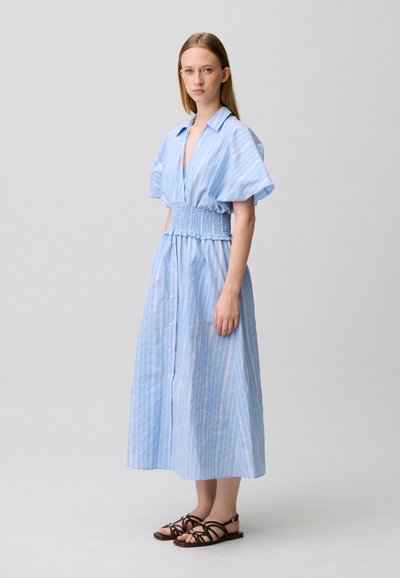 Donna in piedi che indossa un vestito midi azzurro chiaro con maniche corte, bottoni davanti, vita arricciata e sandali neri piatti con cinturini, su uno sfondo neutro.