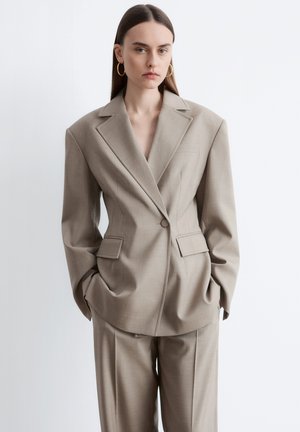 Ung kvinde med langt mørkt hår iført oversized beige jakkesæt med polstrede skuldre og guld øreringe, stående med hænderne i lommerne.