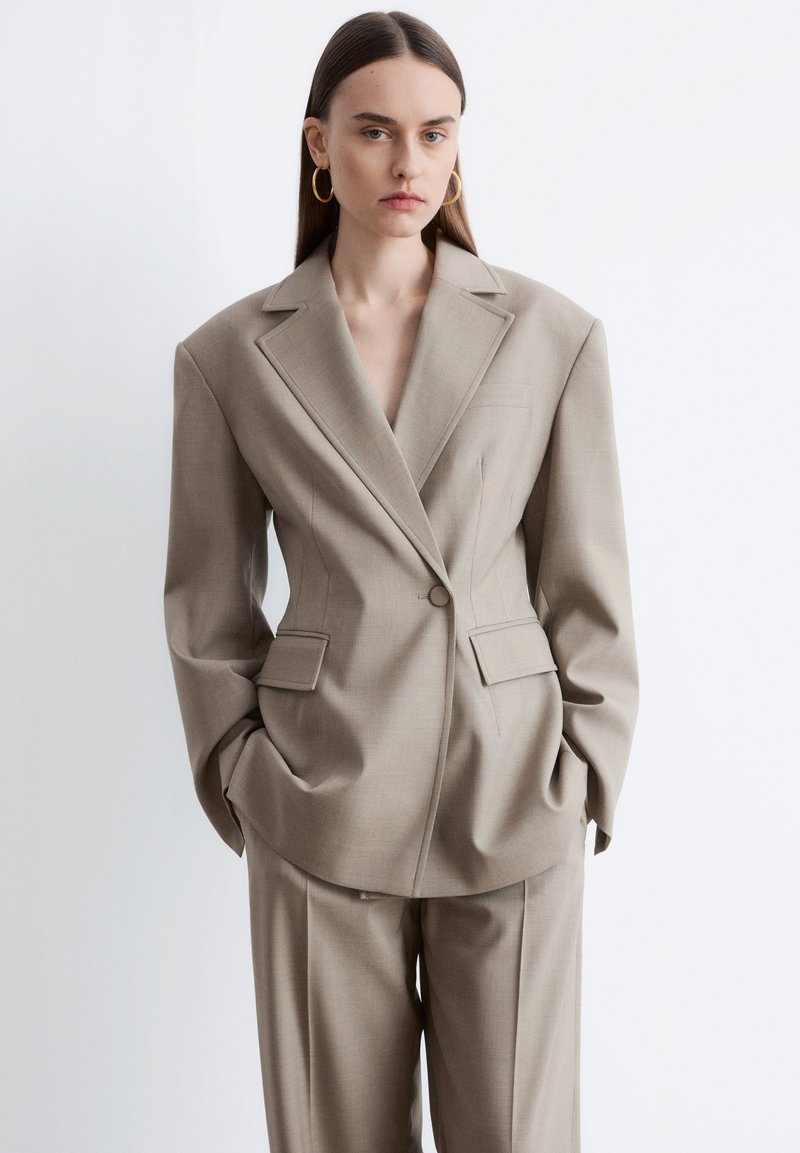 Giovane donna con lunghi capelli scuri che indossa un ampio completo beige con spalle imbottite e orecchini a cerchio dorati, in piedi con le mani nelle tasche.