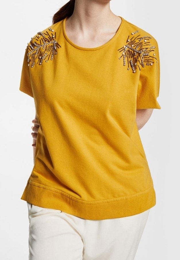 Mujer con camiseta de manga corta de color mostaza con adornos de cuentas en los hombros y pantalones blancos, con las manos detrás de la espalda.