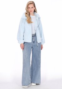Jachetă de fleece albastru deschis, pufoasă, cu glugă, închidere cu fermoar și buzunare laterale, asortată cu un tricou în dungi și blugi largi din denim deschis la culoare.