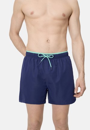 Hombre sin camiseta vistiendo pantalones de baño azul marino con cintura elástica y cordón de color azul claro, de pie contra un fondo liso.