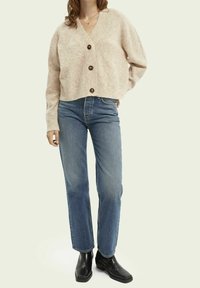 Beige, korte cardigan met geribbelde manchetten en vijf donkere knopen; gecombineerd met blauwe rechte jeans en zwarte enkellaarsjes.
