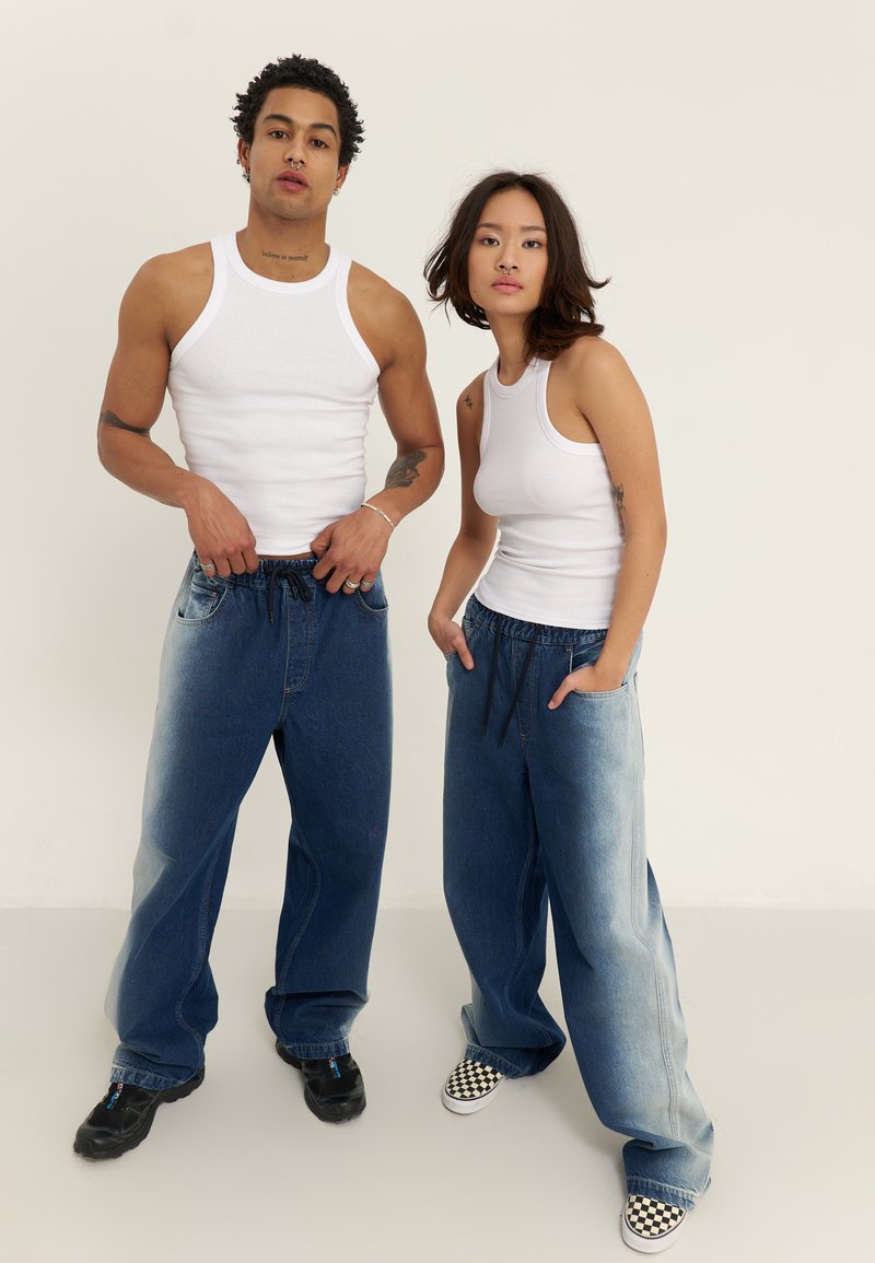 Două modele poartă tricouri albe fără mâneci și pantaloni din denim albastru lejeri cu șnururi. Un model poartă pantofi negri; celălalt are slip-on-uri în carouri.