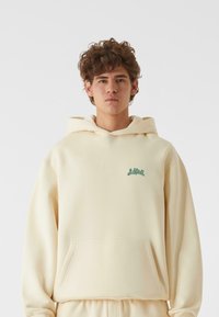 Sudadera color crema hecha de una suave mezcla de algodón, con bolsillo frontal y bordado de logo verde en la parte izquierda del pecho.