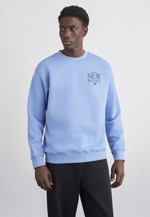 Homme portant un sweat-shirt New Balance bleu clair et un pantalon noir, regardant vers sa gauche sur un fond clair uni.