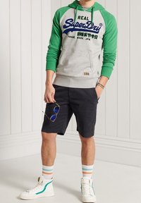 Grijze hoodie met groene mouwen en blauw logo, gecombineerd met donkere shorts, witte sneakers met groene accenten en gestreepte sokken.