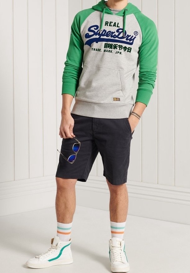 Grijze hoodie met groene mouwen en blauw logo, gecombineerd met donkere shorts, witte sneakers met groene accenten en gestreepte sokken.