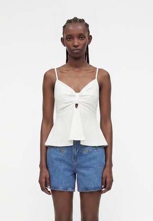 Γυναίκα με πλεγμένα μαλλιά που φοράει λευκό αμάνικο τοπ peplum, συνδυασμένο με ψηλόμεσα μπλε τζιν σορτς με μικρή κεντημένη λουλουδένια διακόσμηση.