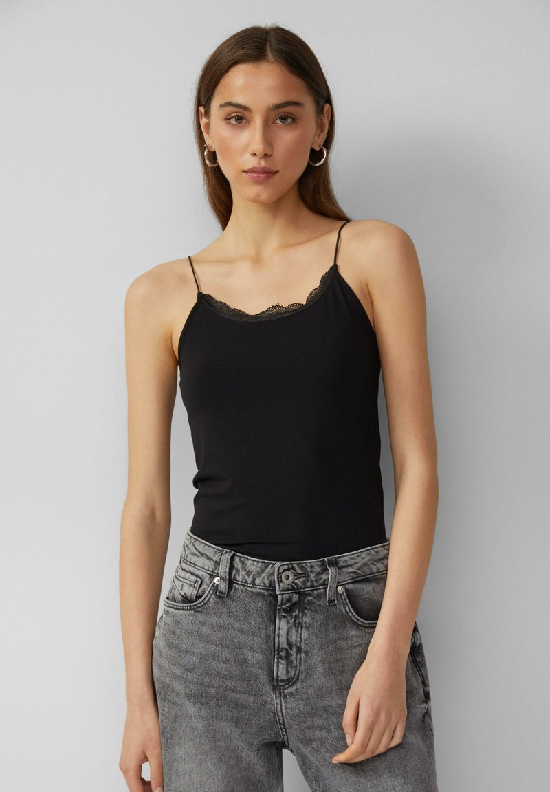 Zwarte cami-top met kantafwerking langs de halslijn, voorzien van dunne verstelbare bandjes, gecombineerd met hooggeknoopte grijze jeans. Gladde stofstructuur.
