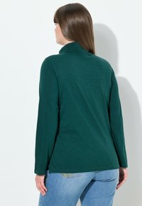 Maglia verde a maniche lunghe con colletto alto, realizzata in tessuto morbido. Accoppiata a jeans di denim azzurri, la vista da dietro mostra una vestibilità rilassata.