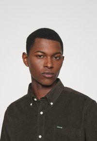 Donkergroene corduroy overhemd met knoopsluiting en zak, met een klein groen Barbour-logo op de borst. Getextureerd stofontwerp.