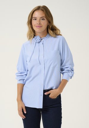 Chemise boutonnée à rayures bleues et blanches avec un col volanté et une fleur en tissu décorative, dotée de manches longues et d'une poche avant.