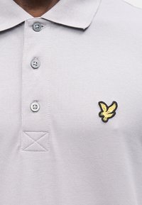 Polo grigio chiaro in tessuto lavorato, con tre bottoni e un logo di un'aquila oro e nero sul petto a sinistra.