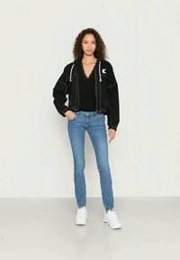 Mulher com cabeloCacheado, vestindo um hoodie preto com zip, uma blusa preta com borda recortada, jeans skinnies azuis e ténis brancos, encostada a uma parede lisa.