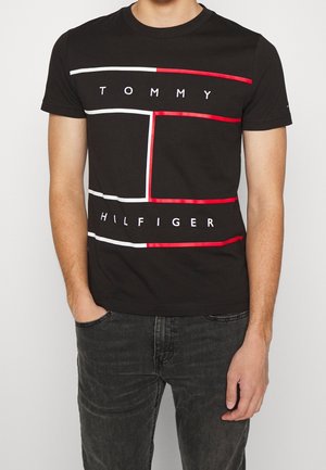 Mies, joka pitää päällään mustaa Tommy Hilfiger -t-paitaa, jossa on punareunainen suorakulmainen kuvio ja brändin nimen teksti edessä.