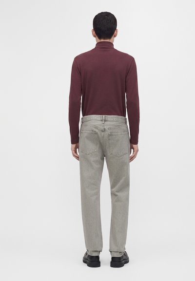 Homme debout de dos, portant un haut à manches longues bordeaux, un jean en denim gris clair et des chaussures noires sur un fond blanc uni.