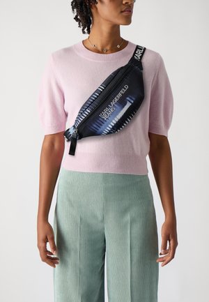 Sort satin slingtaske med et blåt og hvidt grafisk mønster, udstyret med en lynlåslukning og justerbar rem, båret over en lyserød sweater og grønne bukser.