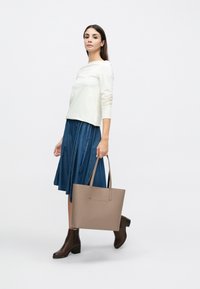 Borsa tote beige con due manici, texture liscia. Modella indossa una maglia color crema e una gonna di denim, camminando con stivaletti marroni alla caviglia. Design semplice e moderno.