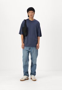 Námořnické oversized tričko a světle modré rovné džíny. Černá crossbody taška, bílé tenisky s gumovou podrážkou. Jednoduché pozadí.
