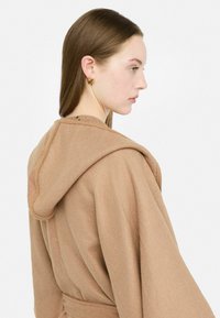 Garbamento marrone con cappuccio e vestibilità rilassata. Realizzato in tessuto morbido, presenta una cintura da annodare e dettagli di cucitura distintivi lungo le spalle.