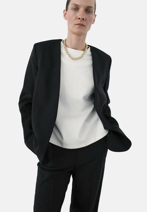 Blazer negro con un diseño estructurado, llevado sobre una blusa blanca. Un collar de cadena de oro texturizado agrega detalle. Ajuste a medida con un aspecto minimalista.