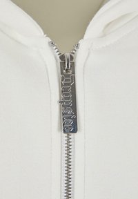 Sweat à fermeture éclair blanche avec une fermeture éclair en métal argenté et une étiquette gravée. Le tissu a une texture douce avec un design simple.