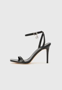 Μαύρο σανδάλι με ψηλό τακούνι, στενό λουράκι στον αστράγαλο, τετράγωνη μύτη, υλικό από λείο δέρμα και μεταλλικό διακοσμητικό πόρπη.