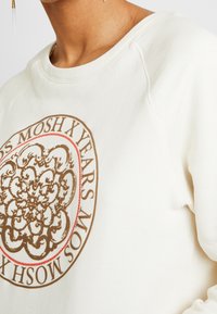 Sudadera de color crema con un diseño gráfico circular en tonos marrón y rojo que incluye el texto "MOSH X YEARS". Tela texturizada con un ajuste relajado y escote redondeado.
