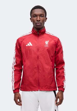 Ung mand iført en rød Liverpool FC Adidas træningsjakke med hvide striber og hvide sportsshorts, der vender fremad mod en ensfarvet baggrund.