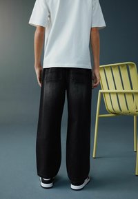 Jeans negri cu picioare largi, croială lejeră și buzunare în spate, asortați cu un tricou alb; purtați cu adidași negri și albi. Un scaun galben în apropiere.